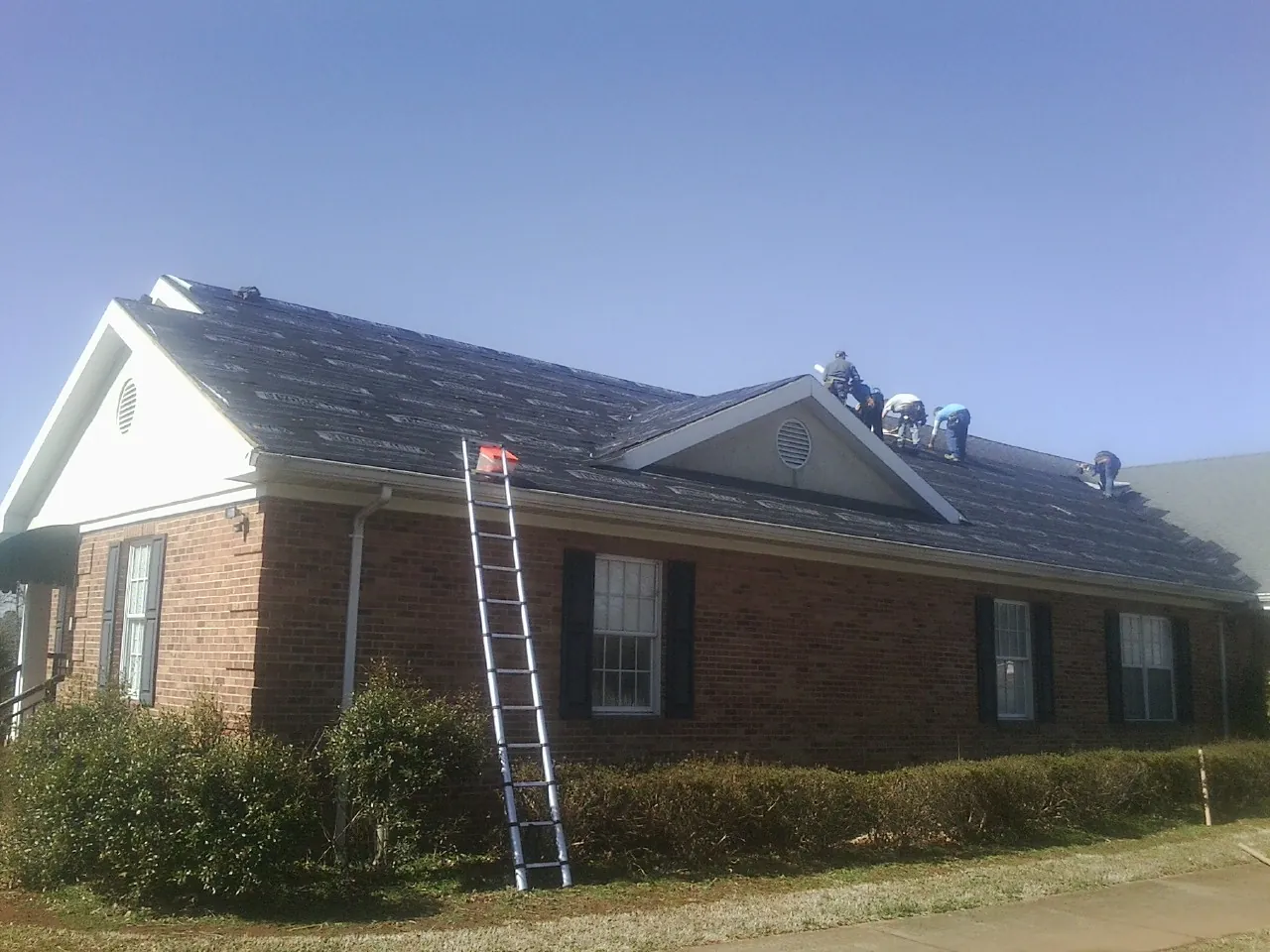 Crew tearing shingles off a brick ranch roof with a ladder set — Emergency Roof Repair work by our crew in Wading River