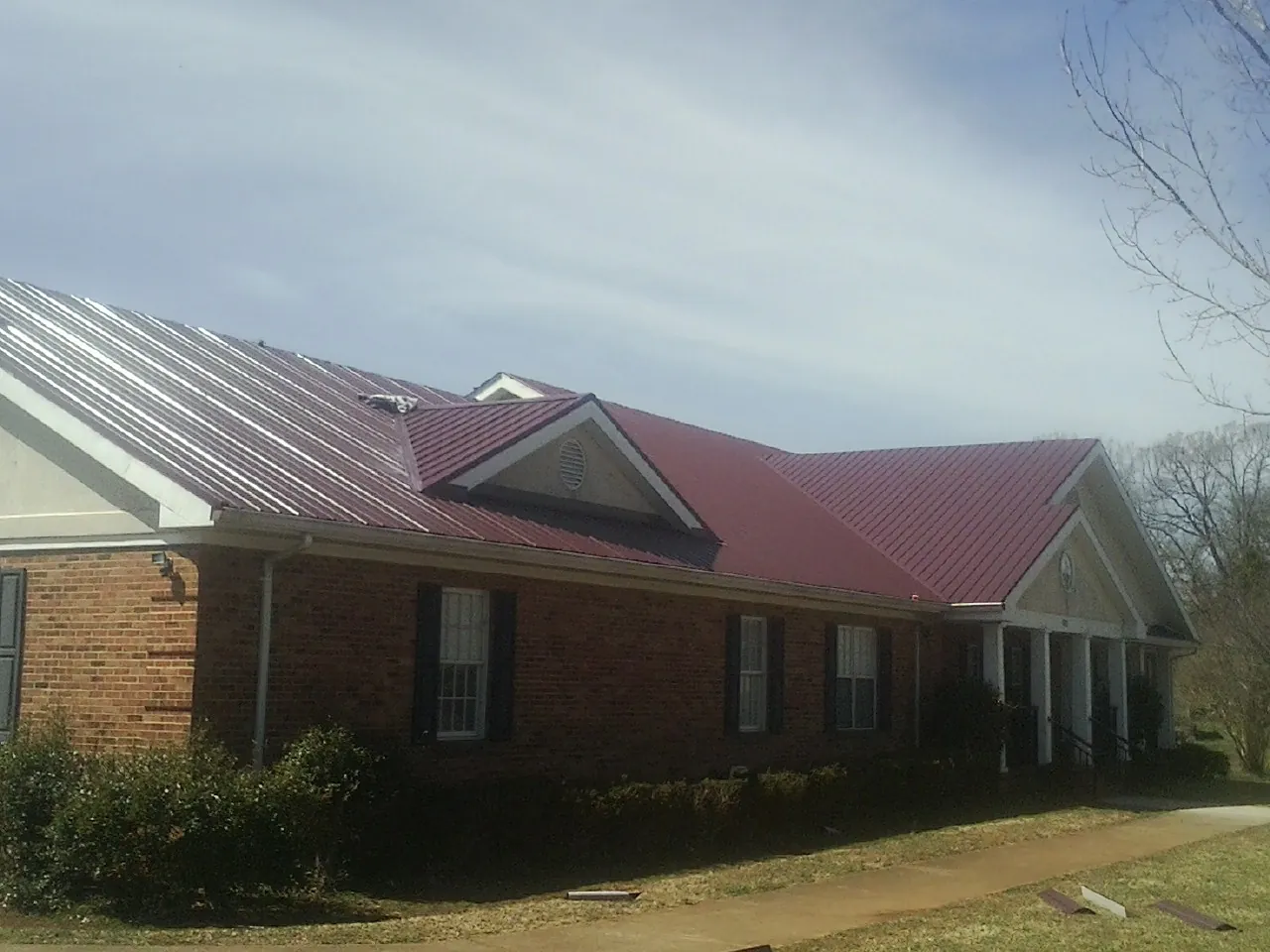 Red metal roof with gabled entries on a long brick building, from a Hail Damage Roof Repair job in Wading River