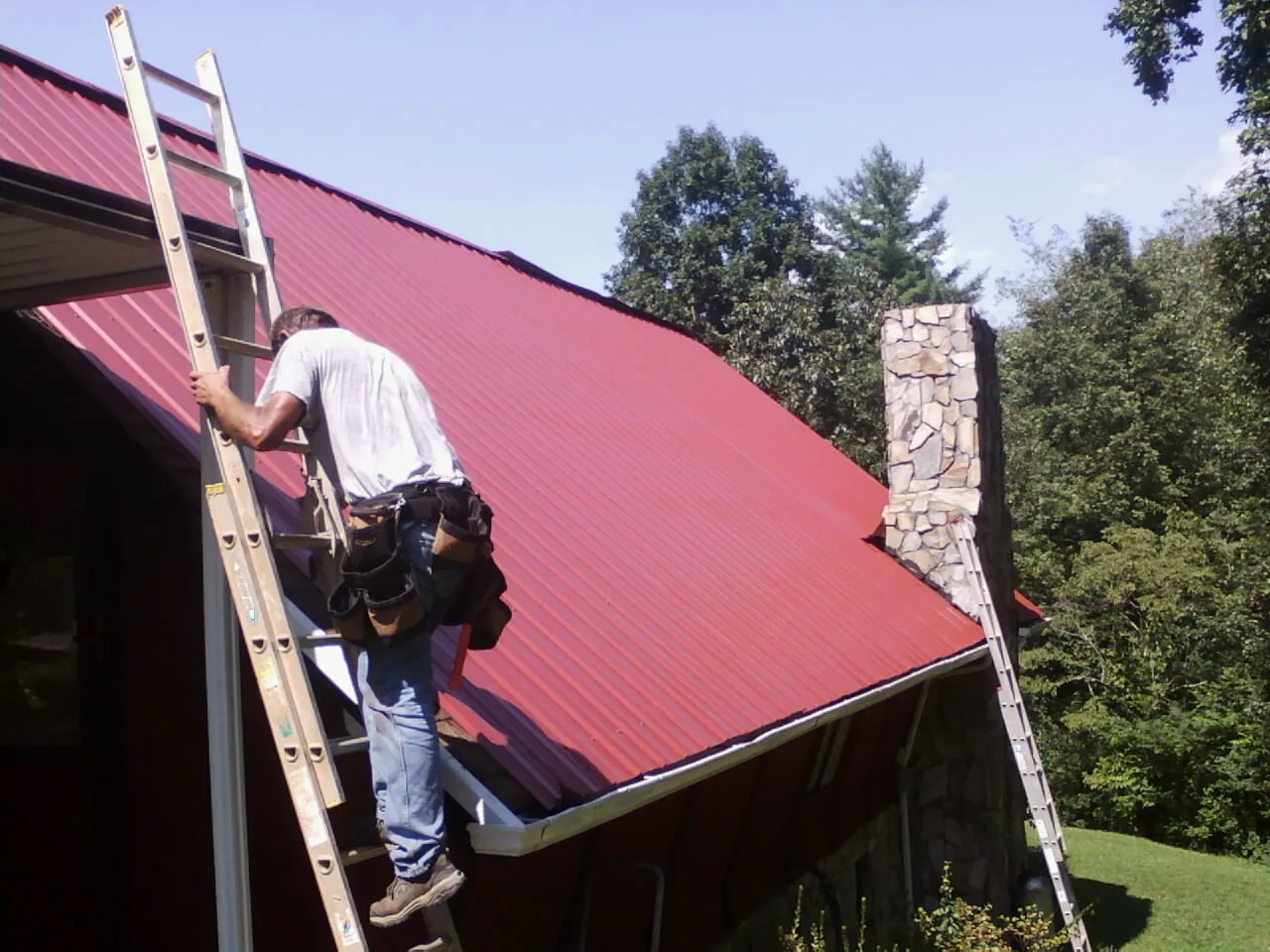 Roofer on a ladder beside a red metal roof and stone chimney — Soffit & Fascia Repair by Roofing Brothers in Wading River